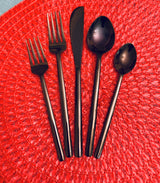 Five pieces of matte black stainless steel flatware, including two forks, a knife, a spoon, and a teaspoon, arranged on a textured red circular placemat.