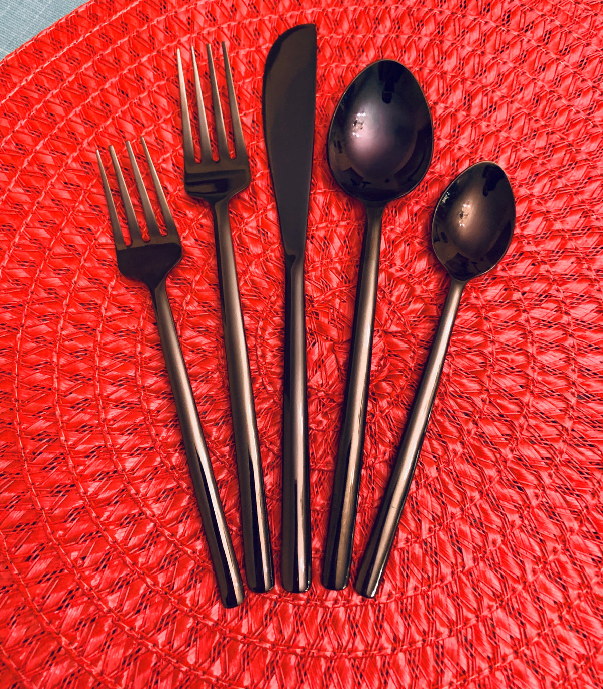 Five pieces of matte black stainless steel flatware, including two forks, a knife, a spoon, and a teaspoon, arranged on a textured red circular placemat.