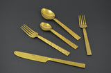 Golden flatware set with textured handles including a knife, two forks, and two spoons arranged on a dark gray surface.