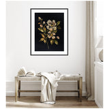 Framed floral art with beige and peach flowers and green leaves on a black background, hung above a woven bench with a draped cream cloth and open book.