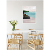 Framed canvas of a seascape with green hills, dark rocks, and calm blue water hanging above a white dining table with light wooden chairs and a glass vase of white calla lilies.
