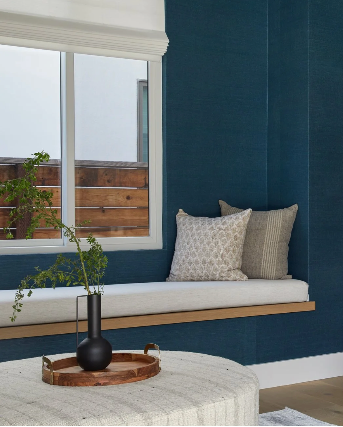 Window seat with a white cushion and two patterned beige pillows against deep blue walls, a round woven ottoman with a wooden tray and black vase with greenery in front.
