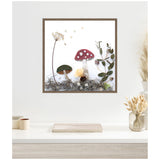 Framed canvas art print depicting mushrooms and botanical elements with a red-capped mushroom having white spots and other greenery, displayed on a white wall above a modern shelf.