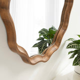 Close-up of a scalloped round mirror with a warm-toned wooden frame showing smooth, flowing curves reflecting soft natural light and green plants nearby.
