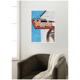 Framed canvas print showing a vintage red and white 'Veggies' sign mounted on a white paneled building against a blue sky background, displayed above a dark olive armchair with a beige knitted throw