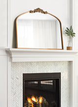 Arched mirror with ornate gold frame and decorative crest on top, positioned above a white mantel with herringbone tile surround and a small plant in a glass vase.
