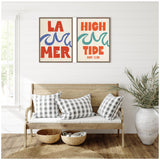 Two framed canvas prints with bold red text and stylized blue and green wave graphics hanging above a wooden bench with three black-and-white checkered pillows in a white room.