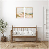 Two pastel landscape paintings with soft blues, greens, and pinks in wooden frames above a wooden bench with white cushions, set against a white wall with a round woven rug.