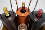 Three cylindrical leather wine totes in dark brown, tan, and black, each holding a bottle with visible necks and cork or foil tops, with embossed logos on top flaps.