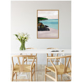 Framed canvas painting of a serene seascape with turquoise water, green cliffs, and dark rocks, hanging on a white wall above a white dining table with wooden chairs and white calla lilies.