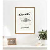 Minimalist framed artwork featuring a sketch of a croissant with handwritten text 'croissant' and 's'il vous plaît' on a light background, hanging above a white countertop with decor.