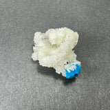 White stilbite mineral cluster with a bright blue cavansite crystal ball attached at the base, displayed on a gray textured surface.