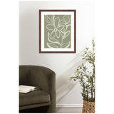 Framed wall art print with white line-drawn delicate flowers and veins on a muted green background, hanging above a dark olive green armchair with an open book and next to a woven basket with a leafy plant.