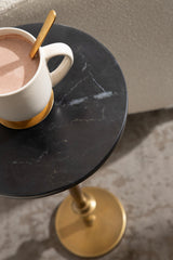 Close-up of a round black marble table top with white veining, holding a white ceramic mug of hot chocolate with a gold spoon, supported by a gold pedestal base.