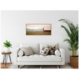 Framed landscape canvas depicting a red barn beside cultivated rows, with distant rolling hills under a soft sky, hanging above a white sofa with patterned pillows.