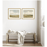 Two framed prints of expansive, muted green and beige meadows under cloudy sky, hung side by side above a wooden bench with a draped white throw and an open book.