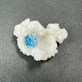 Bright blue cavansite ball nestled within rough, white stilbite crystals on a gray background, showing distinct textures and contrast between the minerals.