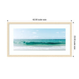 Framed panoramic photo of a calm ocean wave under a blue sky with scattered clouds, bordered by a slim light wood frame and white matting.