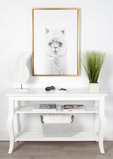 Minimalist white console table with a framed black and white alpaca portrait above, flanked by a white decorative leaf sculpture and green grass plant in a white pot.