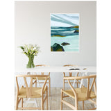 Abstract ocean landscape painting with layered teal, blue, and green brushstrokes above dark rock shapes, framed and hanging above a modern white dining table with wooden chairs and white calla lilies in a vase