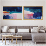 Two abstract canvas paintings with blue, teal, pink, and white tones framed in natural wood, displayed above a gray sectional sofa with striped pillows in a minimalist living room.