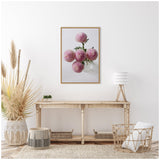 Framed photo of pastel pink dahlia flowers with spherical blooms on a green stem, hanging above a rustic wooden console table in a bright minimalist room.