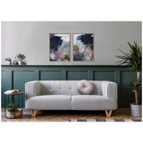 Two framed abstract canvas art pieces with dark blue, gray, pink, and olive tones hang above a light gray mid-century modern sofa with a round pleated gray pillow, against a green paneled wall.