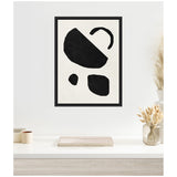 Framed abstract artwork with four irregular black geometric shapes on a white background above a minimalist desk with light wood grain and neutral decor.