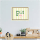 Framed wall art featuring text 'SORRY, I'D RATHER BE playing tennis' with green block letters and pink cursive on pale yellow vertical stripes, hanging above a wooden desk with potted plants and organizer.