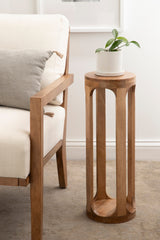 Tall, cylindrical wooden side table with vertical cutouts and rounded edges, holding a small green plant in a white pot beside a cream-colored armchair with a gray pillow.