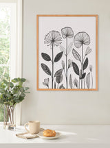 Black and white linocut-style botanical print with tall flowers and patterned leaves in a simple wooden frame hanging on a pale wall above a white table with a plant, cup, and croissant.