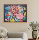 Framed bright coral reef print with pink, red, yellow, and blue coral shapes on a dark background hanging above a light gray sofa with a round cushion and next to a vase of dried flowers on a wooden surface.