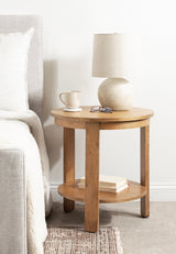 Round two-tier wooden side table with light natural finish, holding a beige ceramic lamp, a white mug, and reading glasses beside a light gray upholstered bed.