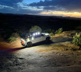 White off-road SUV parked on rocky terrain at dusk with bright roof-mounted LED lights illuminating the ground and nearby bushes under a partly cloudy sky.