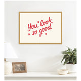 Framed wall art with red handwritten-style text reading 'you look so good' on a white background, hanging above a white shelf with a lamp, photo frame, and potted plant.