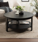 Round black wood coffee table with a lower shelf holding magazines, decorated with a white pot plant and small dark vase on top, placed on a beige rug.
