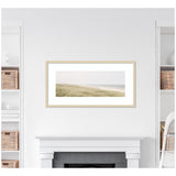 Framed wall art above fireplace showing a serene coastal landscape with grassy dunes and calm sea under an overcast sky.