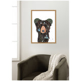 Framed art print of a black bear face made from textured bark, green leaves, and blue flowers on a white background, hanging above a dark olive armchair with a knitted throw.
