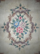 Central floral medallion on a beige wool rug featuring pink, blue, and white flowers surrounded by green leaves and ornate mauve scrollwork.