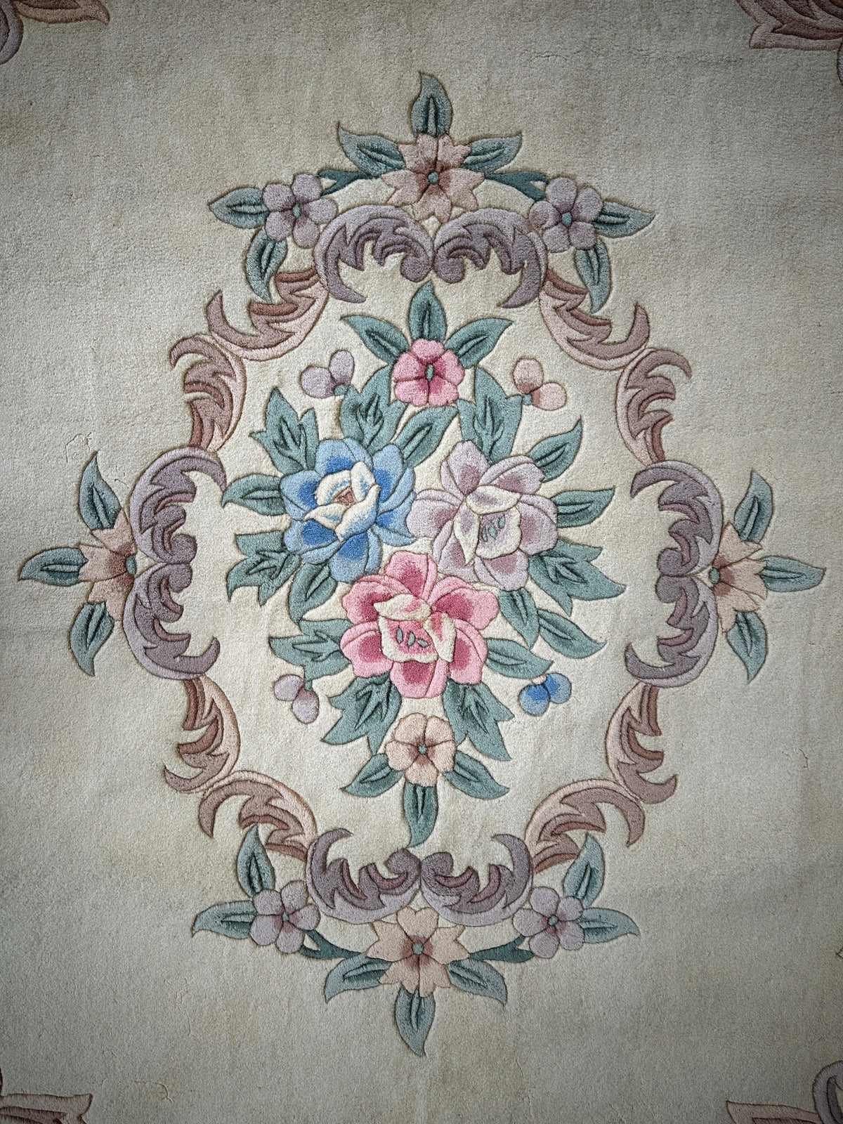 Central floral medallion on a beige wool rug featuring pink, blue, and white flowers surrounded by green leaves and ornate mauve scrollwork.