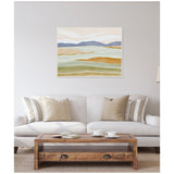 Abstract canvas painting showing layered hills in soft shades of blue, beige, and green above a beige sofa with striped and solid cushions and a wooden coffee table.