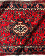Central medallion with geometric patterns on a deep red hand-knotted Persian Hamadan wool rug featuring floral and vine motifs in navy, white, and rust colors.