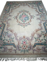 Rectangular hand-tufted wool rug with cream background, central floral medallion, and ornate pastel pink, teal, and beige floral border with fringe edges.