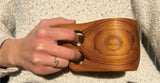 Hand holding a teak kuksa mug featuring smooth wood grain and two circular handle holes, against a textured beige knit background.