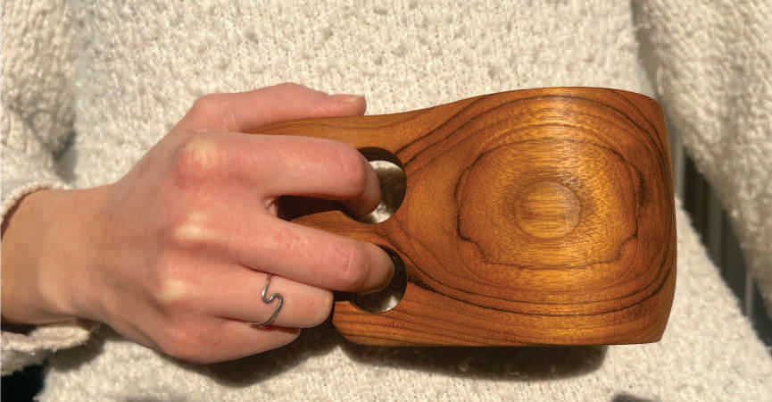 Hand holding a teak kuksa mug featuring smooth wood grain and two circular handle holes, against a textured beige knit background.
