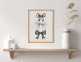 Minimalist wall art with three hand-drawn black bows stacked vertically in varying shading styles within a light wooden frame above a wooden shelf with white vases and dried grasses.