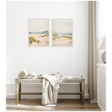 Two soft-toned framed canvas paintings depicting pastel beach landscapes with sand dunes, grassy patches, and distant waters under cloudy skies on a white wall above a woven bench.