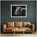 Black and white close-up portrait of a Highland cow with long fur and curved horn, displayed in a black frame on a dark green wall above a brown leather sofa.