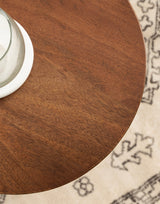 Close-up of a round Dahl wood drink table with a smooth brown wood grain surface, partially showing a glass on a white coaster, over a beige rug with black geometric patterns.