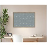 Blue pinboard with a repeating white botanical block print pattern, framed in natural wood, hanging above a minimalist desk with books and a plant nearby.